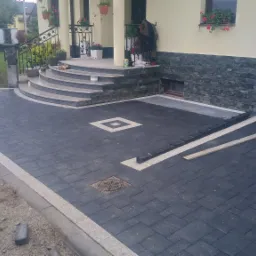 Ułożona ciemnoszara kostka brukowa z jasną obwódką i dekoracyjnymi elementami przed domem z kamiennymi schodami i ozdobnymi kwiatami w donicach.