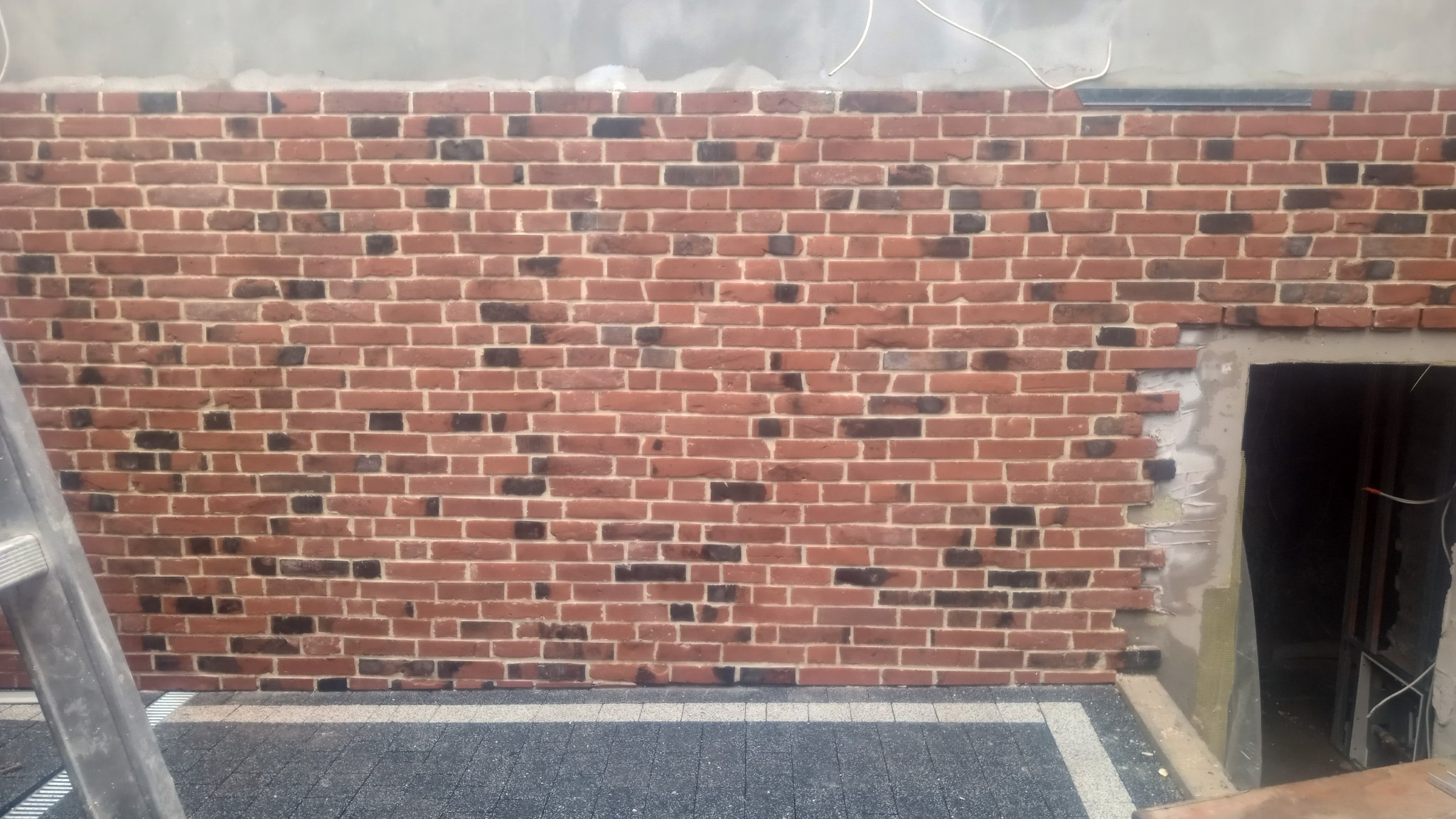 Ceglana ściana z czerwono-czarnych cegieł w trakcie budowy, widoczna dziura w murze obok której leży drabina, a pod ścianą ułożona kostka brukowa z szarych elementów.