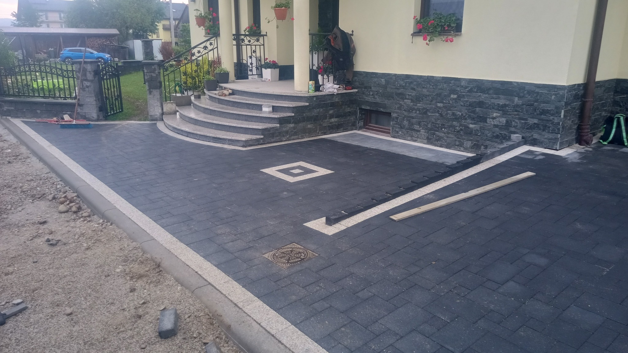 Ułożona ciemnoszara kostka brukowa z jasną obwódką i dekoracyjnymi elementami przed domem z kamiennymi schodami i ozdobnymi kwiatami w donicach.