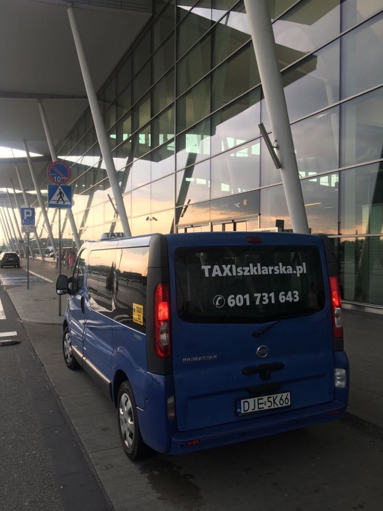 Niebieski van z napisem TAXIszklarska.pl i numerem telefonu zaparkowany przed nowoczesnym budynkiem lotniska o zmierzchu.