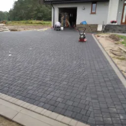 Ułożona, ciemnoszara kostka brukowa na podjeździe przed domem z szarym obrzeżem, widoczny pracownik doglądający powierzchni i ubijarka wibracyjna, w tle garaż.
