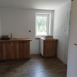 Kamant - Wnętrze kuchni w trakcie prac wykończeniowych z drewnianymi szafkami, brązową podłogą imitującą drewno i oknem z widokiem na zieleń.