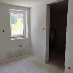 Kamant - Pokój w trakcie prac przygotowawczych do wykończenia, widoczne ślady po szpachlowaniu ścian, wystające kable elektryczne i rury hydrauliczne, surowy betonowy sufit i podłoga, okno z widokiem...