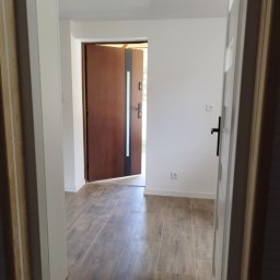 Kamant - Wykończony korytarz z drewnopodobną podłogą, białe ściany z czarnymi włącznikami, brązowe drzwi wejściowe z czarnymi elementami dekoracyjnymi.