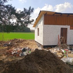 Kamant - Surowy stan budowy domu jednorodzinnego z białych bloczków, widoczne ślady prac ziemnych, gruz i drabina oparta o ścianę, w tle zielone pole i las.