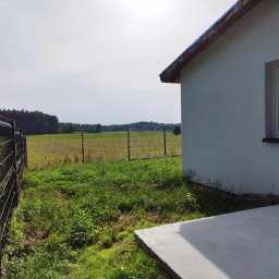 Kamant - Nowy dom z oknem i świeżo wylany betonowy taras, widok na okoliczne pola i las, ogrodzenie panelowe w tle.