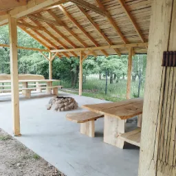 Zadaszona wiata z drewnianymi stołami i ławkami o surowym wykończeniu, miejsce na ognisko z kamieni oraz zestaw szpikulców do pieczenia zawieszony na ścianie.