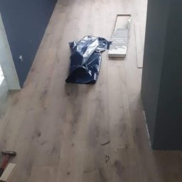 Kamant - Układanie paneli podłogowych w pokoju z ciemną ścianą, widoczny młotek, paczka paneli i worek na odpady.