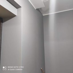 Kamant - Świeżo pomalowane na szaro ściany w narożniku pomieszczenia, widoczne listwy przysufitowe i fragment drewnianego kija w dolnym rogu.