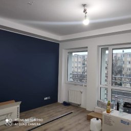 Kamant - Świeżo pomalowane na granatowo ściany w jasnym pokoju z drewnianą podłogą, widoczne okna z widokiem na blok, grzejnik i kartony z akcesoriami budowlanymi.