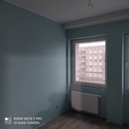 Kamant - Pomieszczenie po malowaniu ścian na kolor turkusowy, widoczne okno z widokiem na blok, grzejnik i fragment podłogi z panelami.