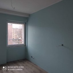 Kamant - Świeżo pomalowane na błękitno ściany pokoju z oknem, widoczne wystające kable elektryczne i gniazdka, podłoga wyłożona panelami imitującymi drewno.