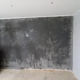 Pracownik w Skierniewicach aplikuje dekoracyjną, ciemnoszarą powłokę na ścianę w pomieszczeniu z surowym podłożem, widoczne puszki elektryczne.