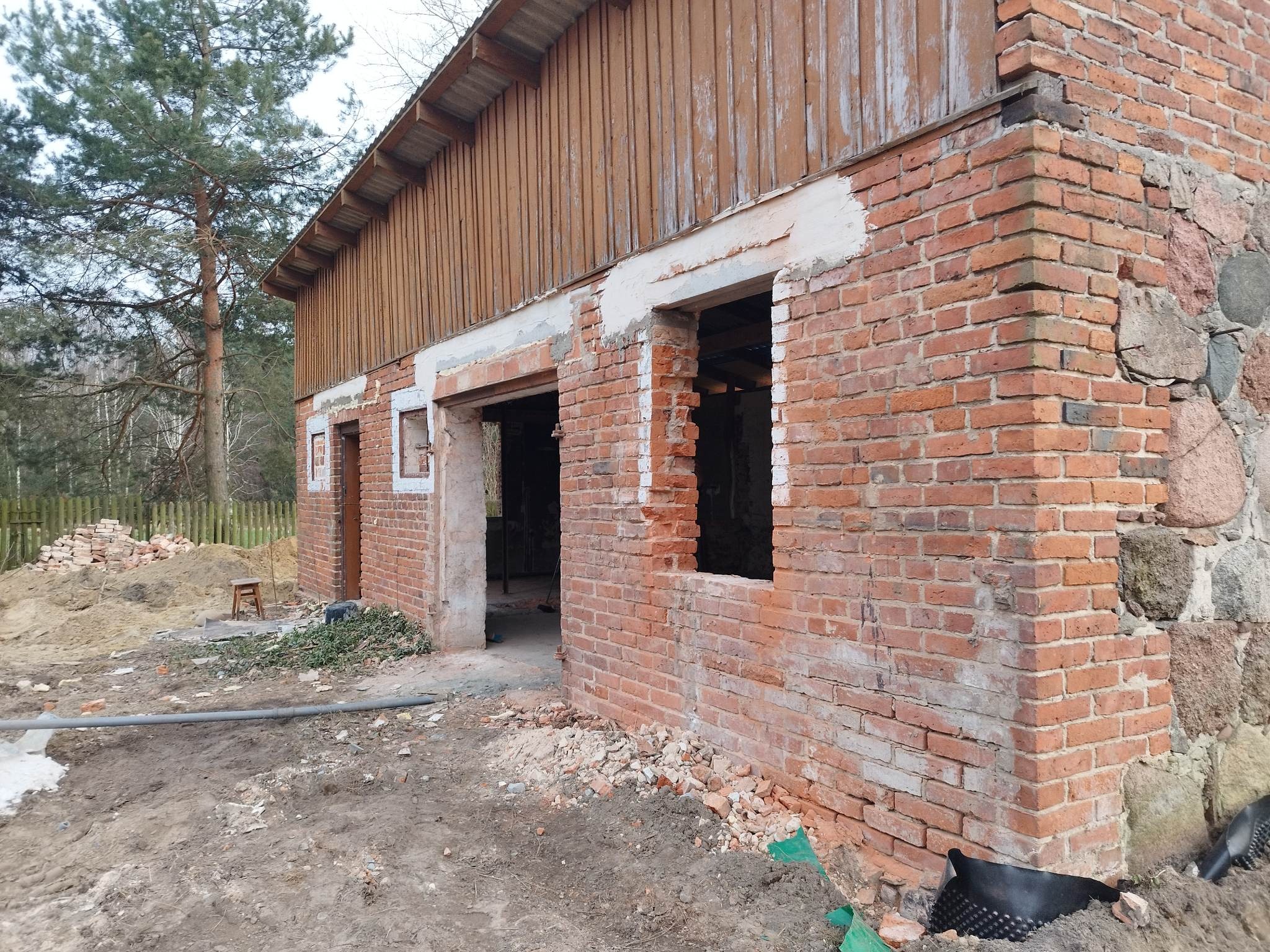 Ceglany budynek w trakcie renowacji z widocznymi otworami okiennymi i drzwiowymi, z częściowo odnowioną elewacją i stertą gruzu na ziemi.