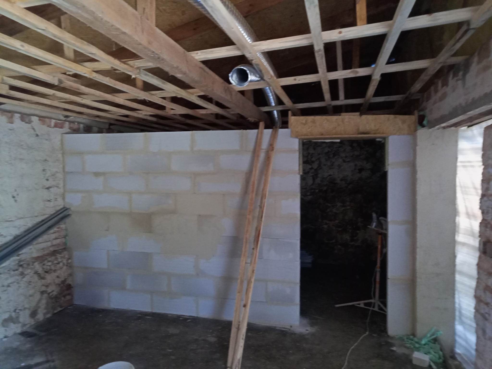Wnętrze w trakcie budowy: ściana z bloczków betonowych, drewniany szkielet sufitu z widoczną rurą wentylacyjną, fragment ściany z cegły i drewniana drabina oparta o ścianę z bloczków.