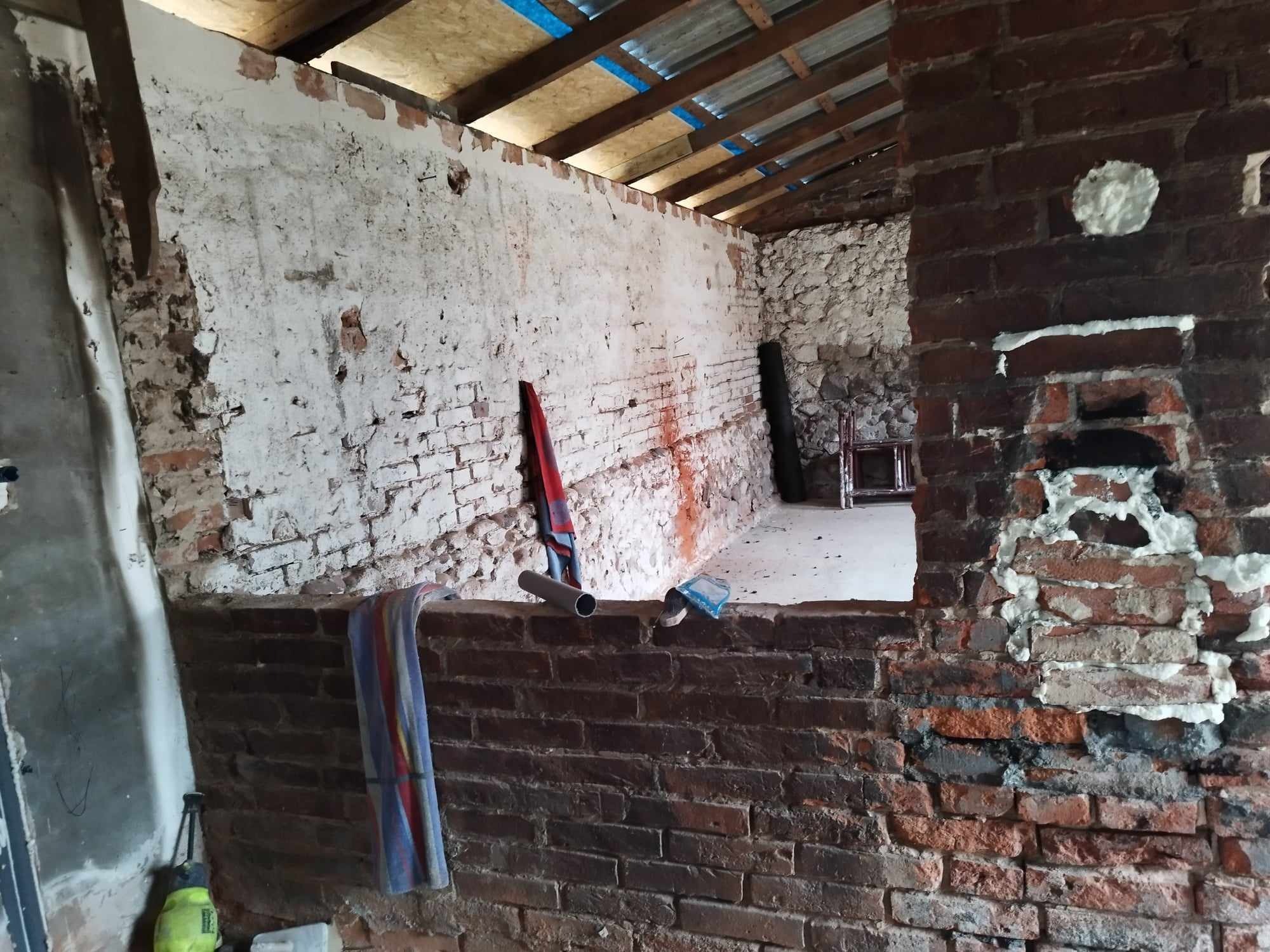 Wnętrze w trakcie prac przygotowawczych do remontu: surowe ceglane ściany z pozostałościami tynku, widoczne narzędzia i materiały budowlane, odsłonięta konstrukcja dachu z izolacją.