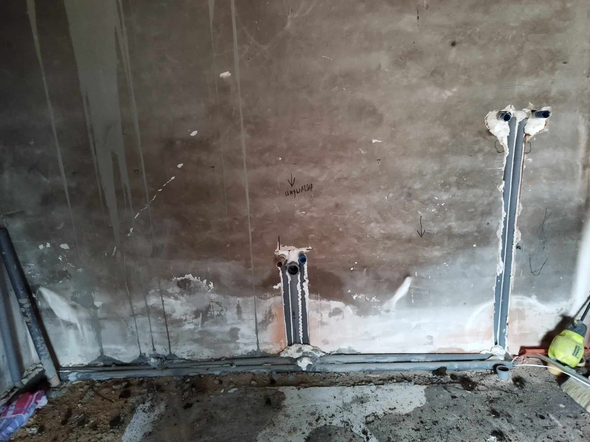 Surowa ściana z widocznymi rurami instalacji wodnej i kanalizacyjnej, zabezpieczonymi pianką montażową, z napisem 'umywalka' nad jednym z otworów, oraz śladami po wcześniejszych pracach...