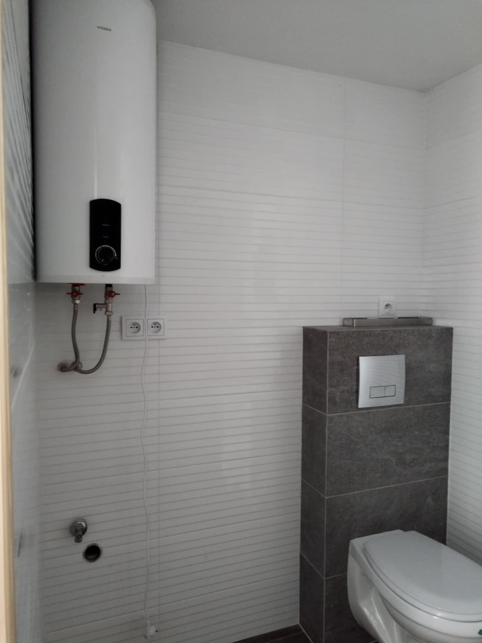 Nowoczesna łazienka z białymi płytkami imitującymi drewno, podwieszana toaleta z szarą obudową z płytek oraz biały bojler elektryczny na ścianie.