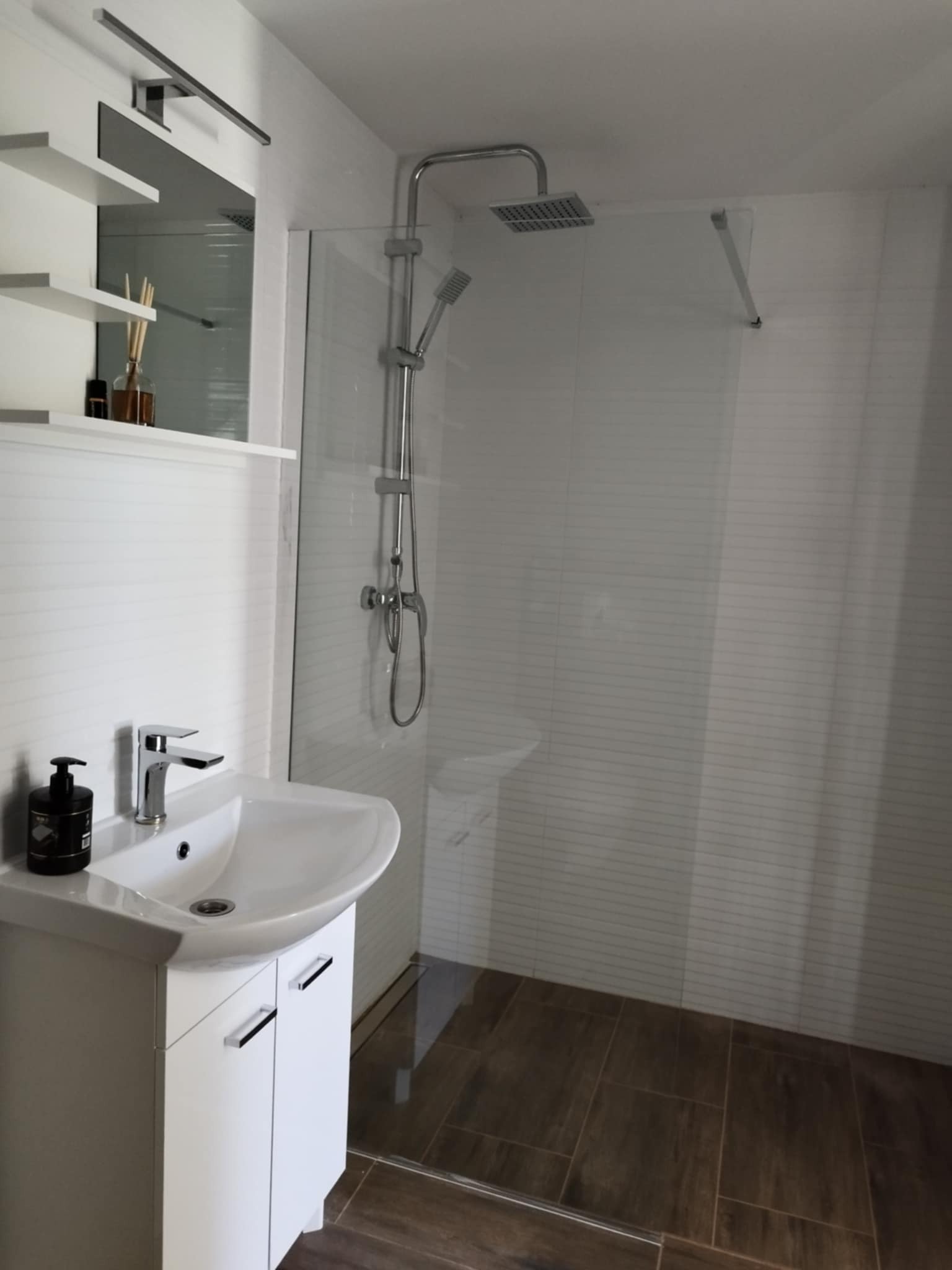 Nowoczesna łazienka z białą umywalką nablatową, kabiną prysznicową typu walk-in z deszczownicą i brązowymi płytkami podłogowymi imitującymi drewno.