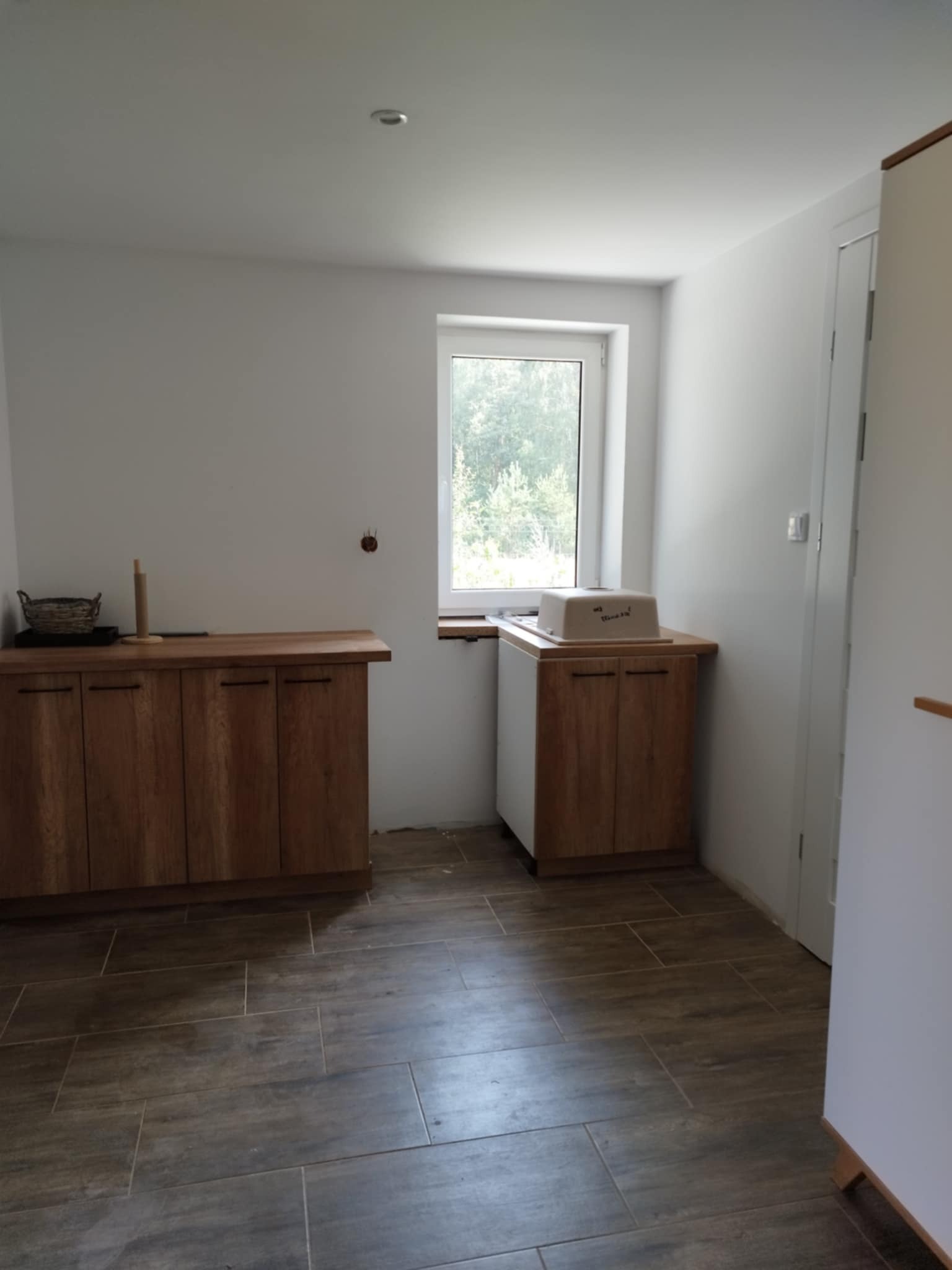 Wnętrze kuchni w trakcie prac wykończeniowych z drewnianymi szafkami, brązową podłogą imitującą drewno i oknem z widokiem na zieleń.