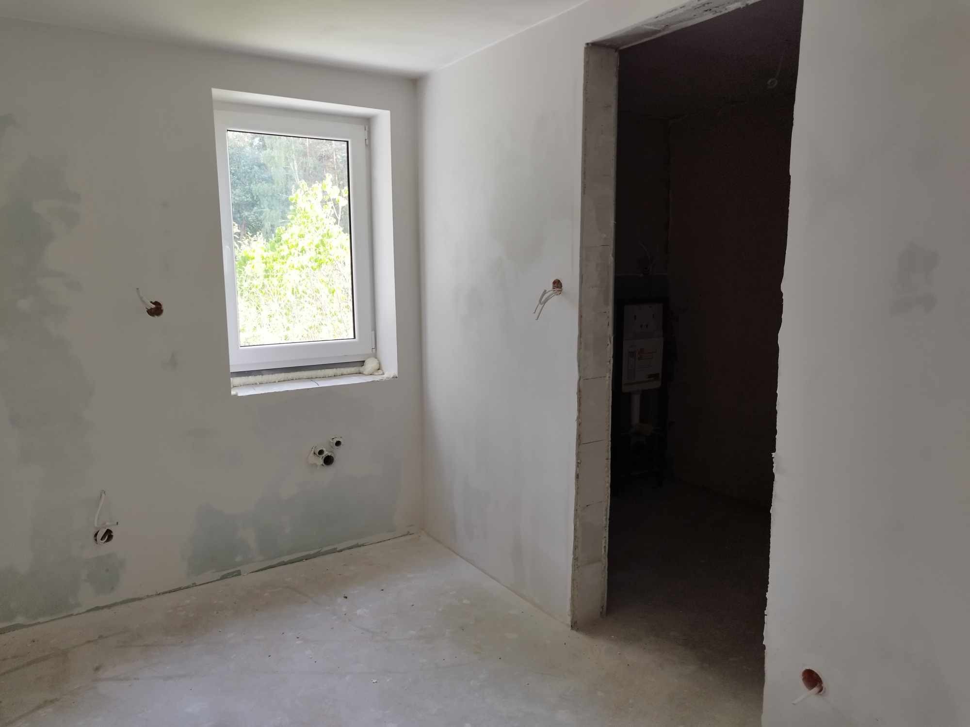 Pokój w trakcie prac przygotowawczych do wykończenia, widoczne ślady po szpachlowaniu ścian, wystające kable elektryczne i rury hydrauliczne, surowy betonowy sufit i podłoga, okno z widokiem...