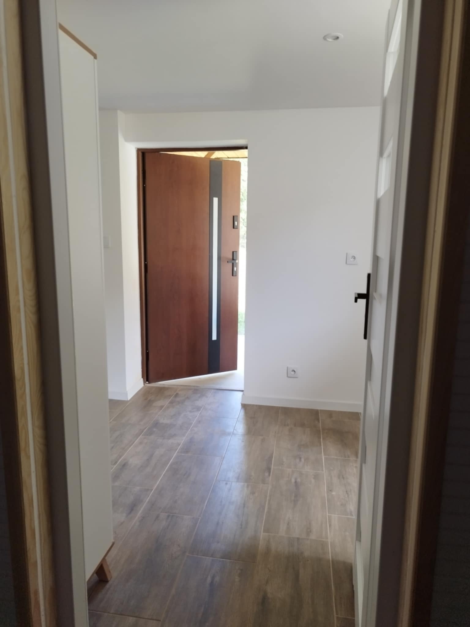 Wykończony korytarz z drewnopodobną podłogą, białe ściany z czarnymi włącznikami, brązowe drzwi wejściowe z czarnymi elementami dekoracyjnymi.