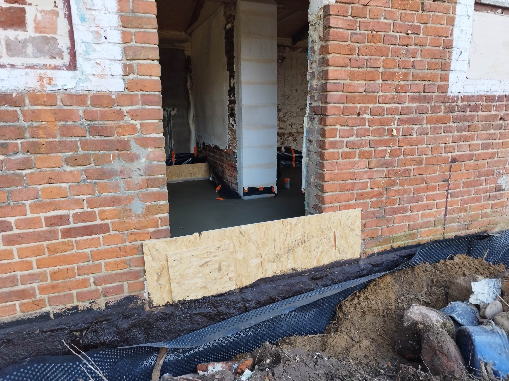 Wnętrze remontowanego budynku z surową cegłą, widoczna izolacja fundamentów z folii kubełkowej i wylewka betonowa wewnątrz.