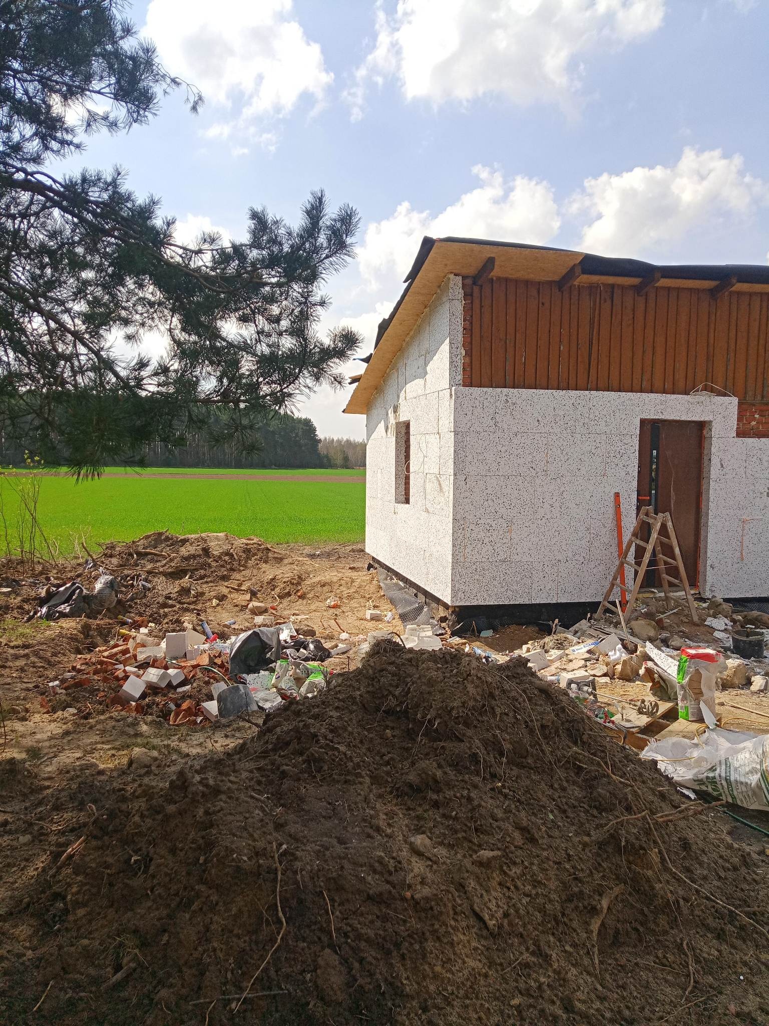 Surowy stan budowy domu jednorodzinnego z białych bloczków, widoczne ślady prac ziemnych, gruz i drabina oparta o ścianę, w tle zielone pole i las.