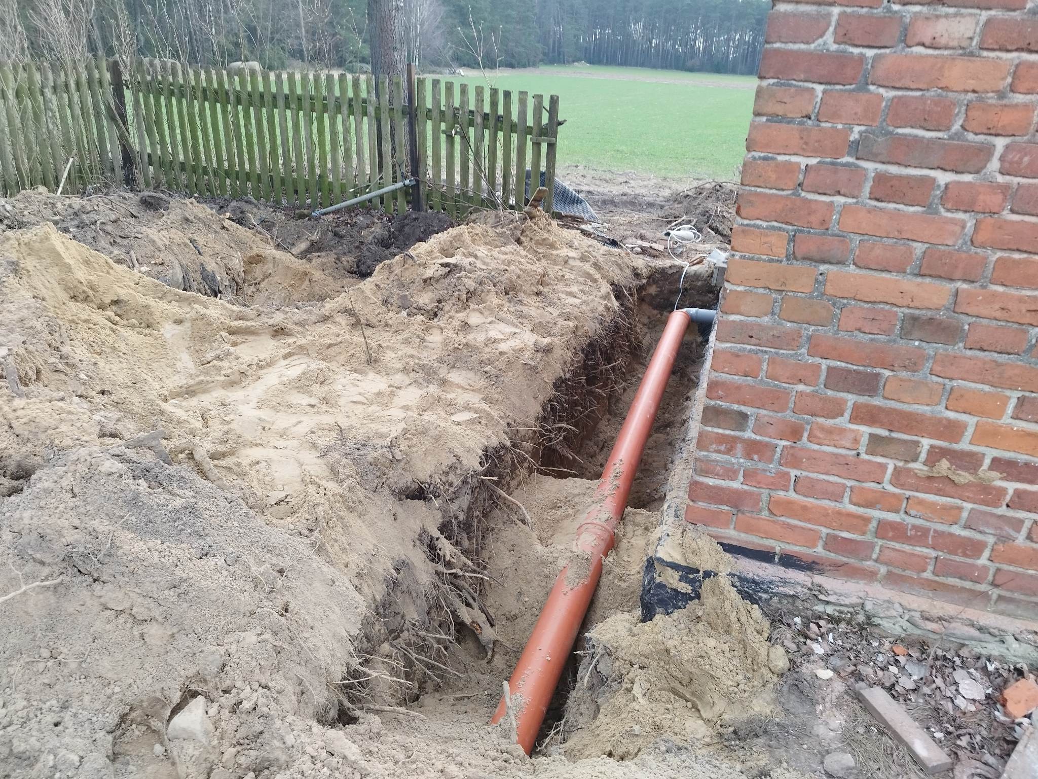 Pomarańczowa rura kanalizacyjna ułożona w wykopie przy ceglanej ścianie budynku, widoczny fragment drewnianego płotu i pole w tle.