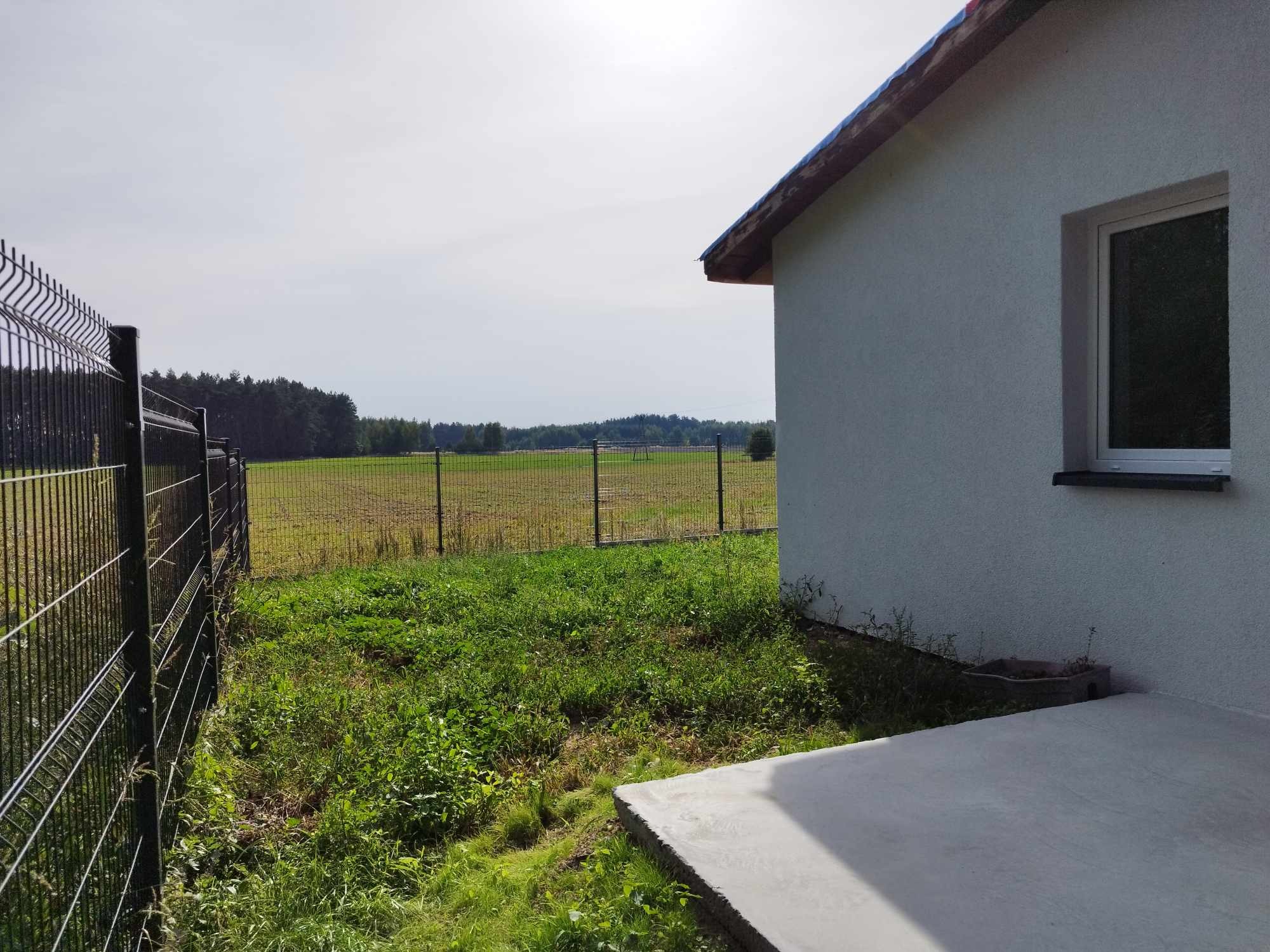 Nowy dom z oknem i świeżo wylany betonowy taras, widok na okoliczne pola i las, ogrodzenie panelowe w tle.