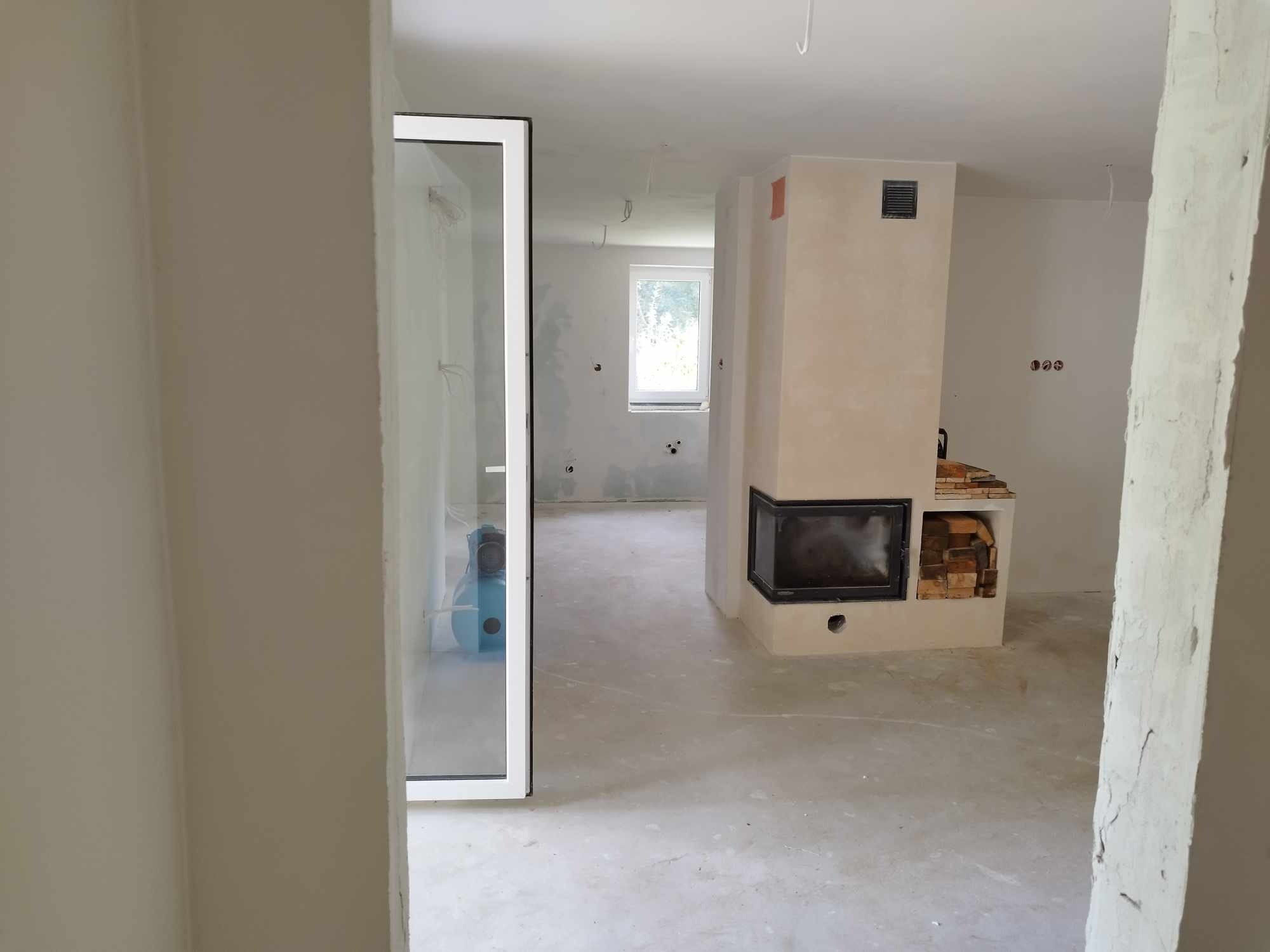 Wnętrze w trakcie prac wykończeniowych: widoczny kominek z obudową, surowa podłoga, ściany przygotowane pod malowanie oraz niebieski agregat malarski za szklanymi drzwiami.