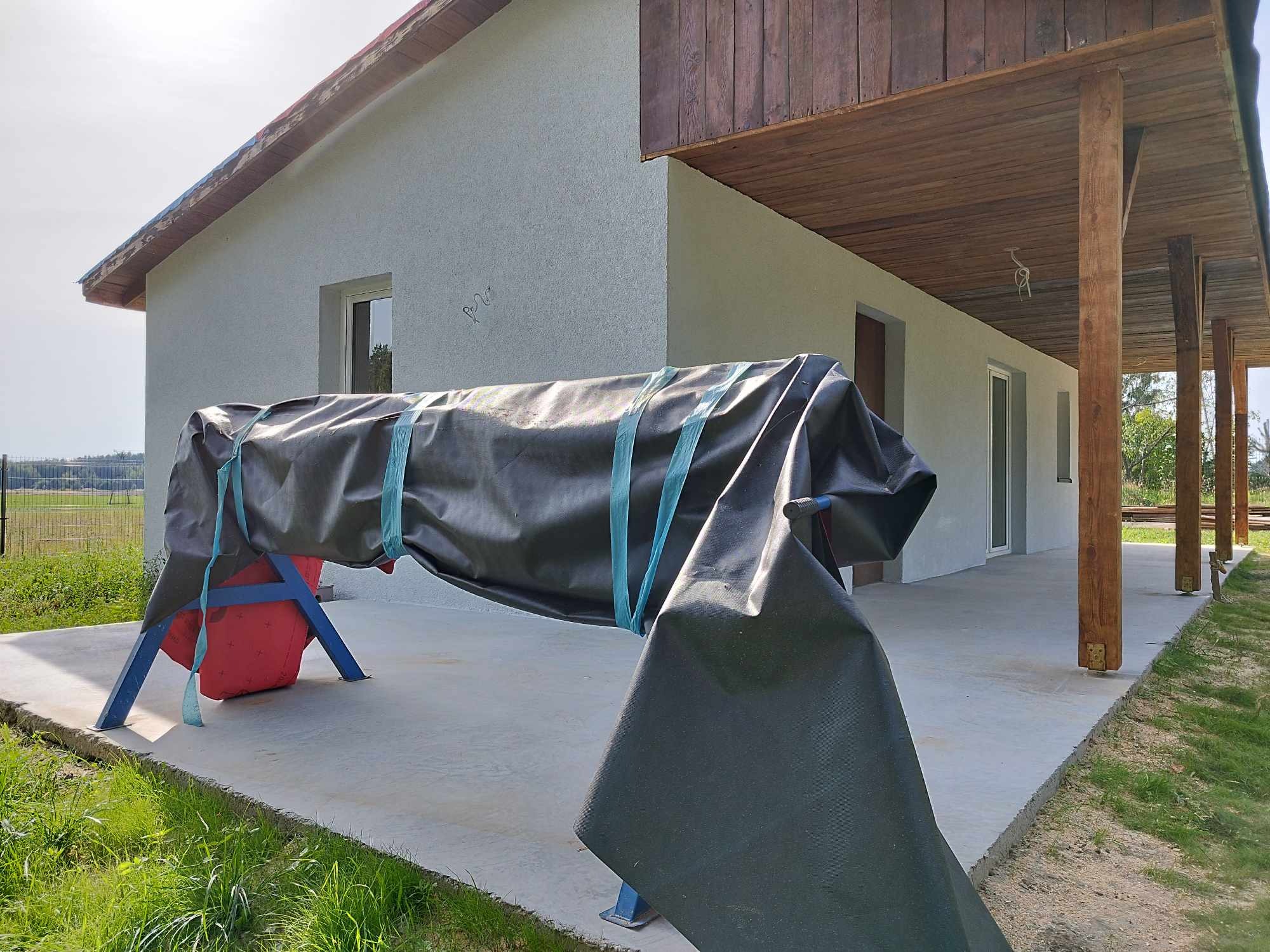 Elewacja domu z szarą strukturą, częściowo zakryta czarną plandeką na niebieskim stelażu, z widocznym fragmentem podbitki drewnianej i kolumn werandy.