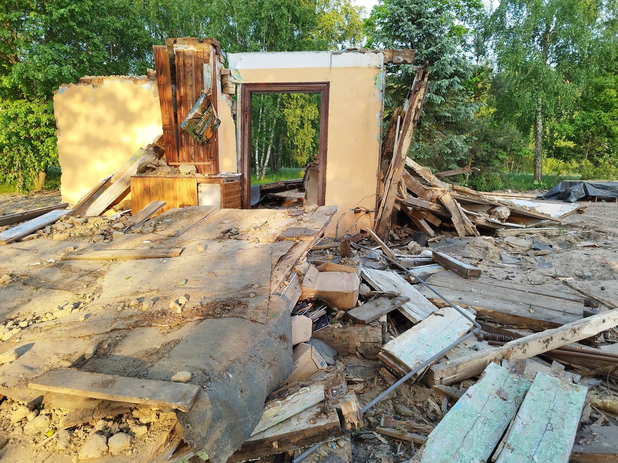 Zrujnowany budynek z widocznymi fragmentami ścian, drewnianymi elementami konstrukcyjnymi, gruzem i otworami okiennymi, na tle zieleni drzew.