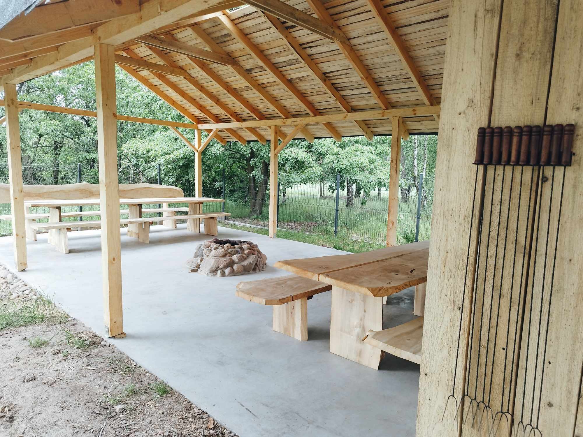 Zadaszona wiata z drewnianymi stołami i ławkami o surowym wykończeniu, miejsce na ognisko z kamieni oraz zestaw szpikulców do pieczenia zawieszony na ścianie.