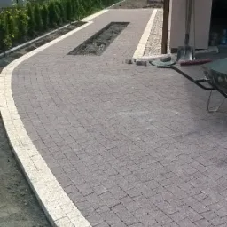 Układanie szarej kostki brukowej z jasnymi obrzeżami, widoczny fragment rabaty, narzędzia budowlane i taczka w tle.