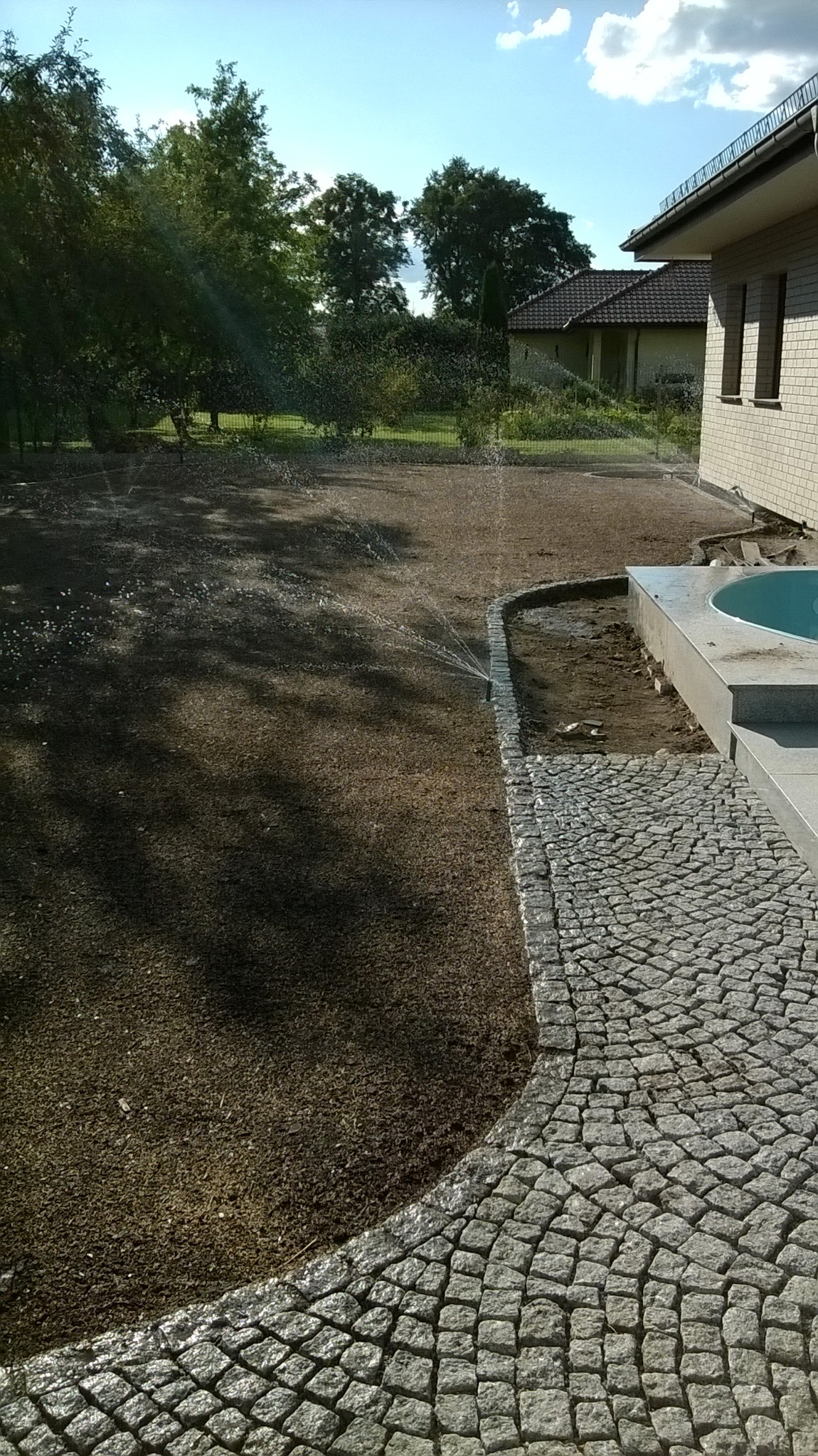 Automatyczne nawadnianie trawnika z systemem zraszaczy, obrzeże z kostki granitowej, widok na basen i dom w tle.