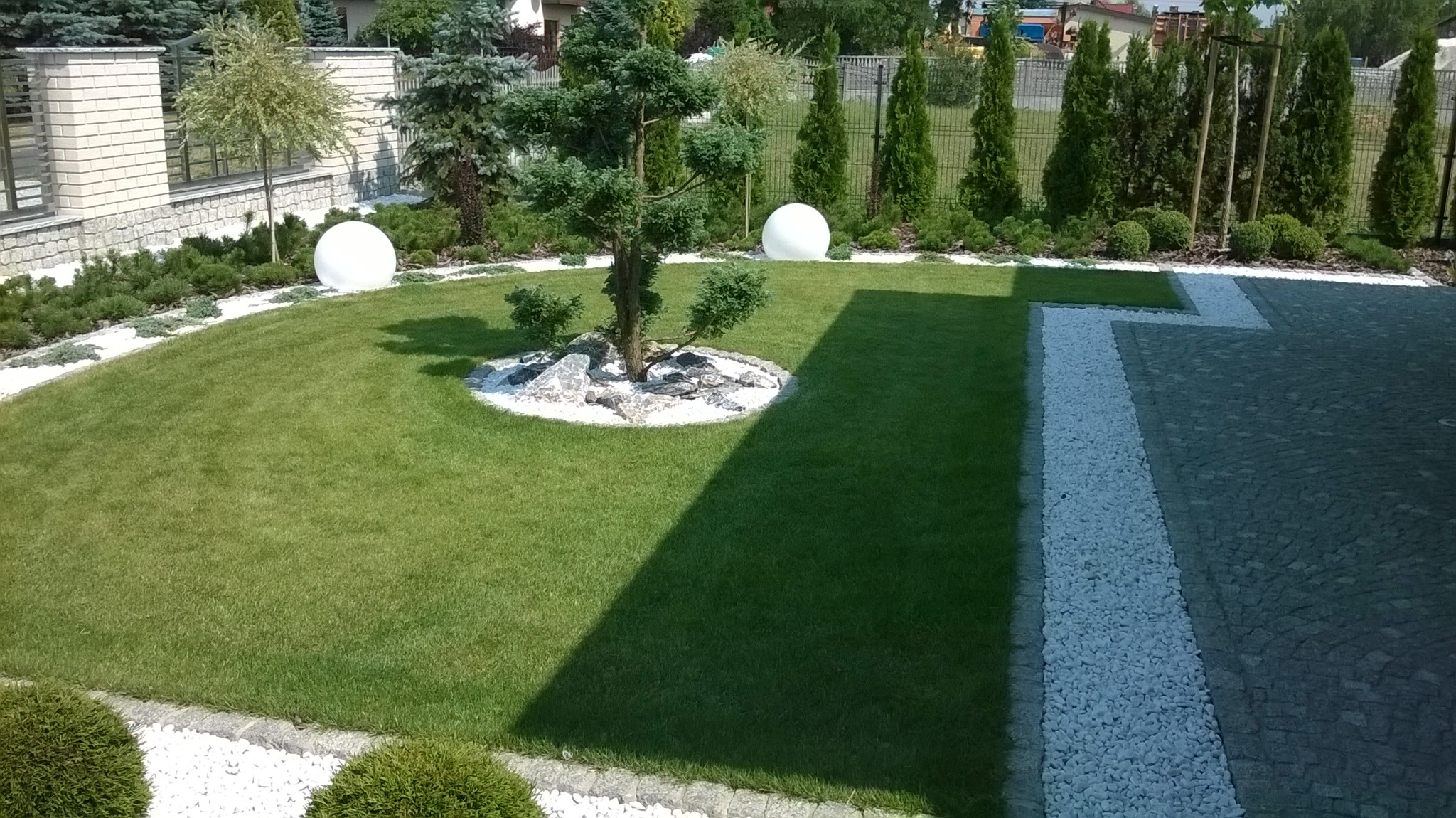 Zadbany trawnik z ozdobnym drzewem bonsai otoczonym białymi kamieniami, kuliste lampy ogrodowe, wykończenie z białego żwiru i kostka brukowa w tle, regularne nasadzenia drzew i krzewów wzdłuż...