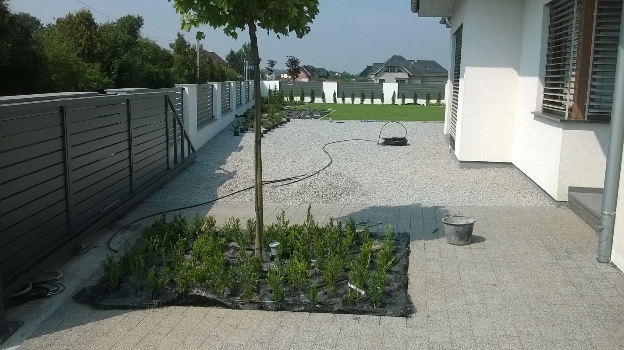 Przygotowany teren pod nasadzenia w ogrodzie: młode krzewy na czarnej tkaninie, żwirowe podłoże, widoczny system nawadniania, nowoczesne ogrodzenie panelowe i fragment domu.
