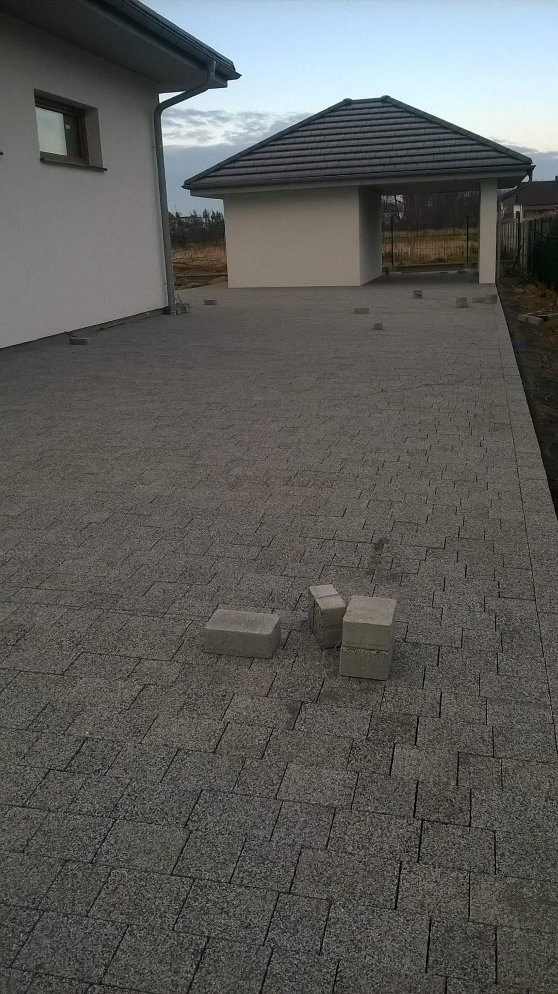 Fragment podjazdu z ułożonej szarej kostki granitowej. W oddali widoczny dom i wiata garażowa. Na pierwszym planie nieułożone kostki.
