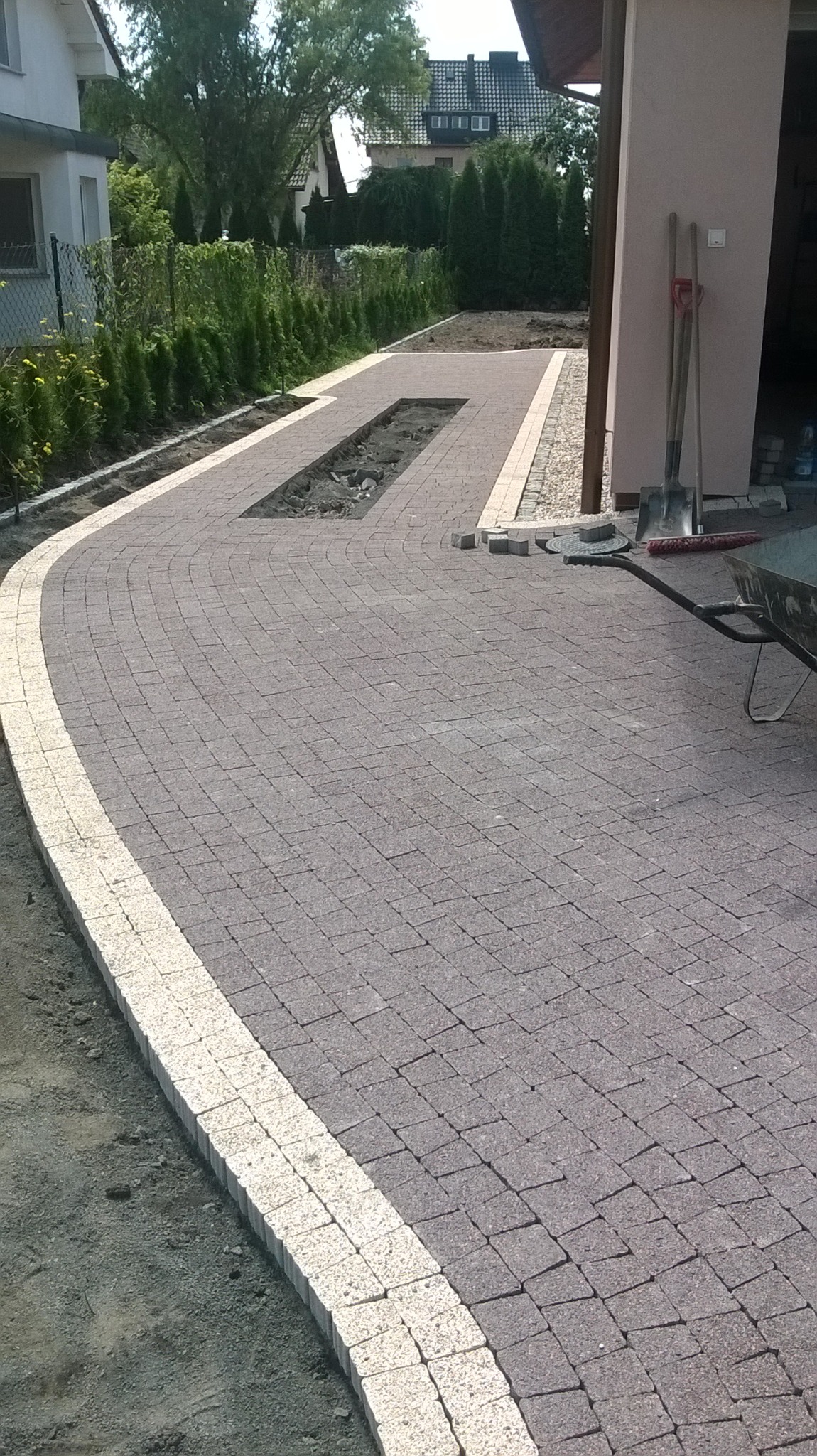 Układanie szarej kostki brukowej z jasnymi obrzeżami, widoczny fragment rabaty, narzędzia budowlane i taczka w tle.