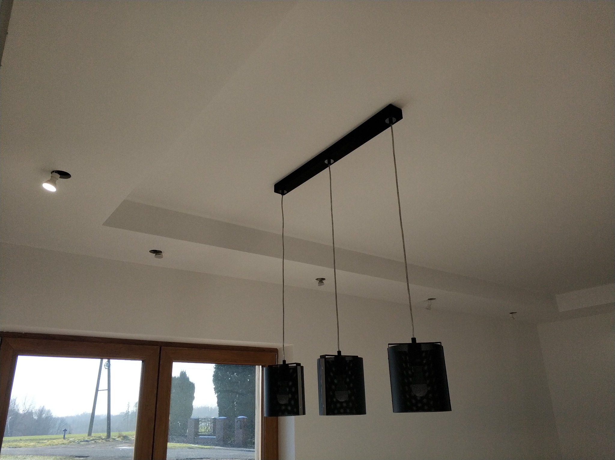 Biały sufit z wbudowanym oświetleniem punktowym i czarną, podłużną lampą wiszącą z trzema abażurami, widok na okno z drewnianą ramą i krajobraz za nim.