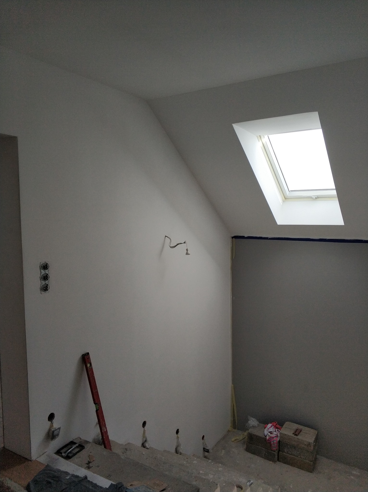 Poddasze w trakcie remontu: świeżo pomalowane ściany na biało, okno dachowe, widoczne puszki elektryczne, poziomica oparta o ścianę, zabezpieczona taśmą granica malowania.