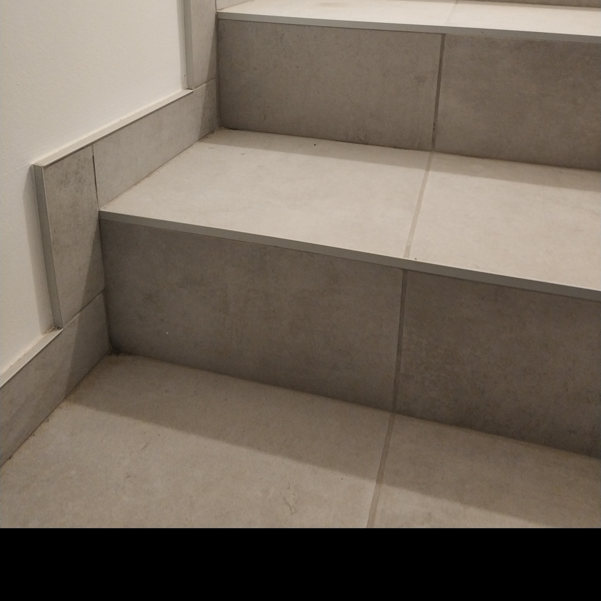 Fragment schodów wykończonych płytkami w odcieniach szarości, z widocznymi fugami i listwami przypodłogowymi z tego samego materiału.