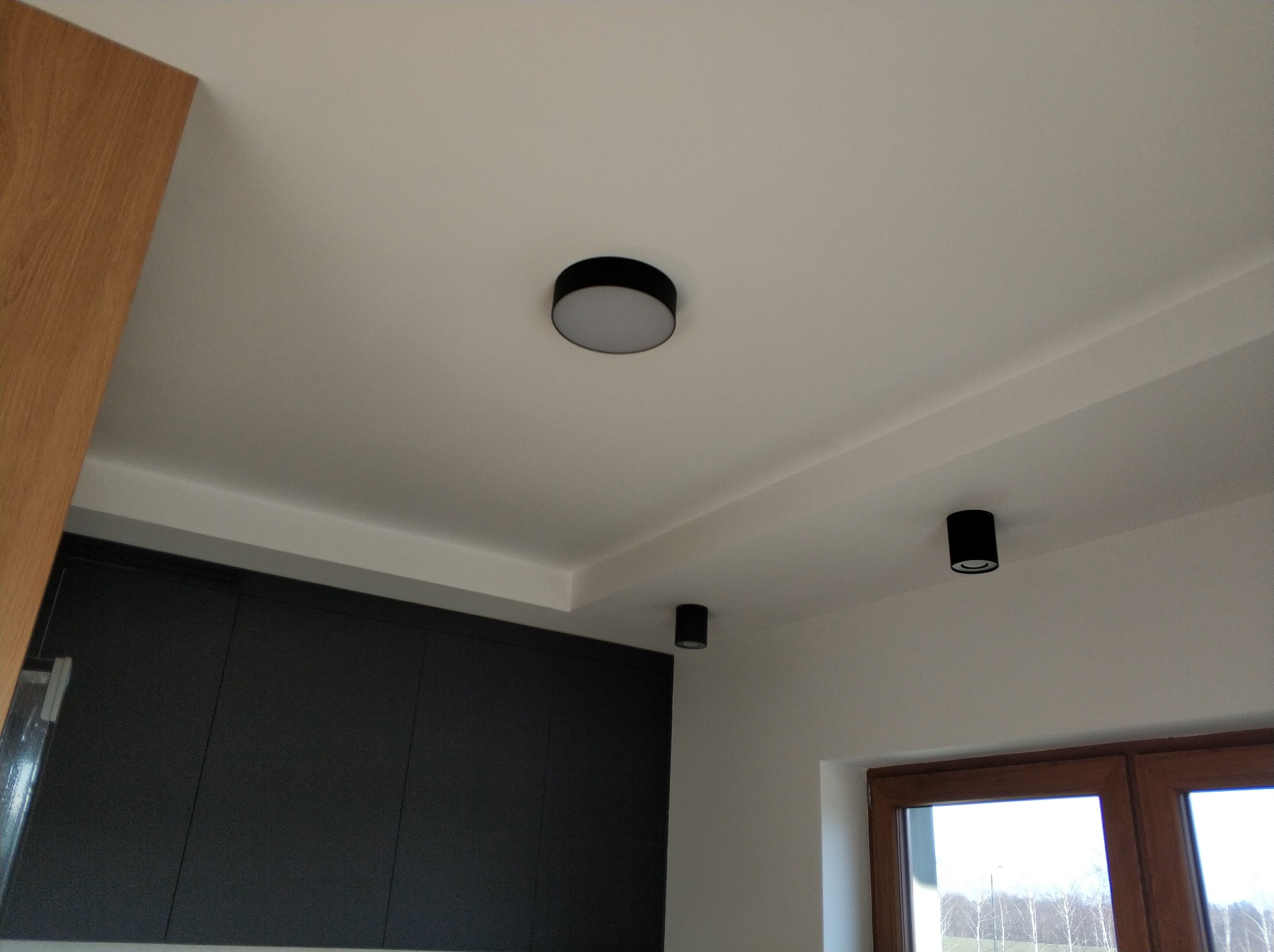 Wnętrze po remoncie w Wodzisławiu Śląskim z widocznymi nowymi, czarnymi lampami sufitowymi, gładkimi ścianami i fragmentem zabudowy kuchennej w ciemnym kolorze.