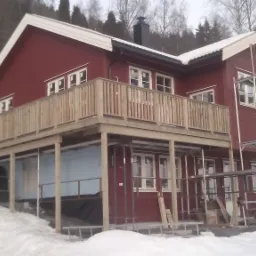 Czerwony dom z drewnianym balkonem na podwyższeniu, w trakcie budowy lub wykończenia, otoczony śniegiem, widoczne rusztowania i materiały budowlane.