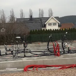 Fundamenty domu z rozprowadzonymi instalacjami: czarne rury kanalizacyjne i czerwone rury osłonowe kabli elektrycznych, widoczne również elementy szalunków i otoczenie działki z domami.