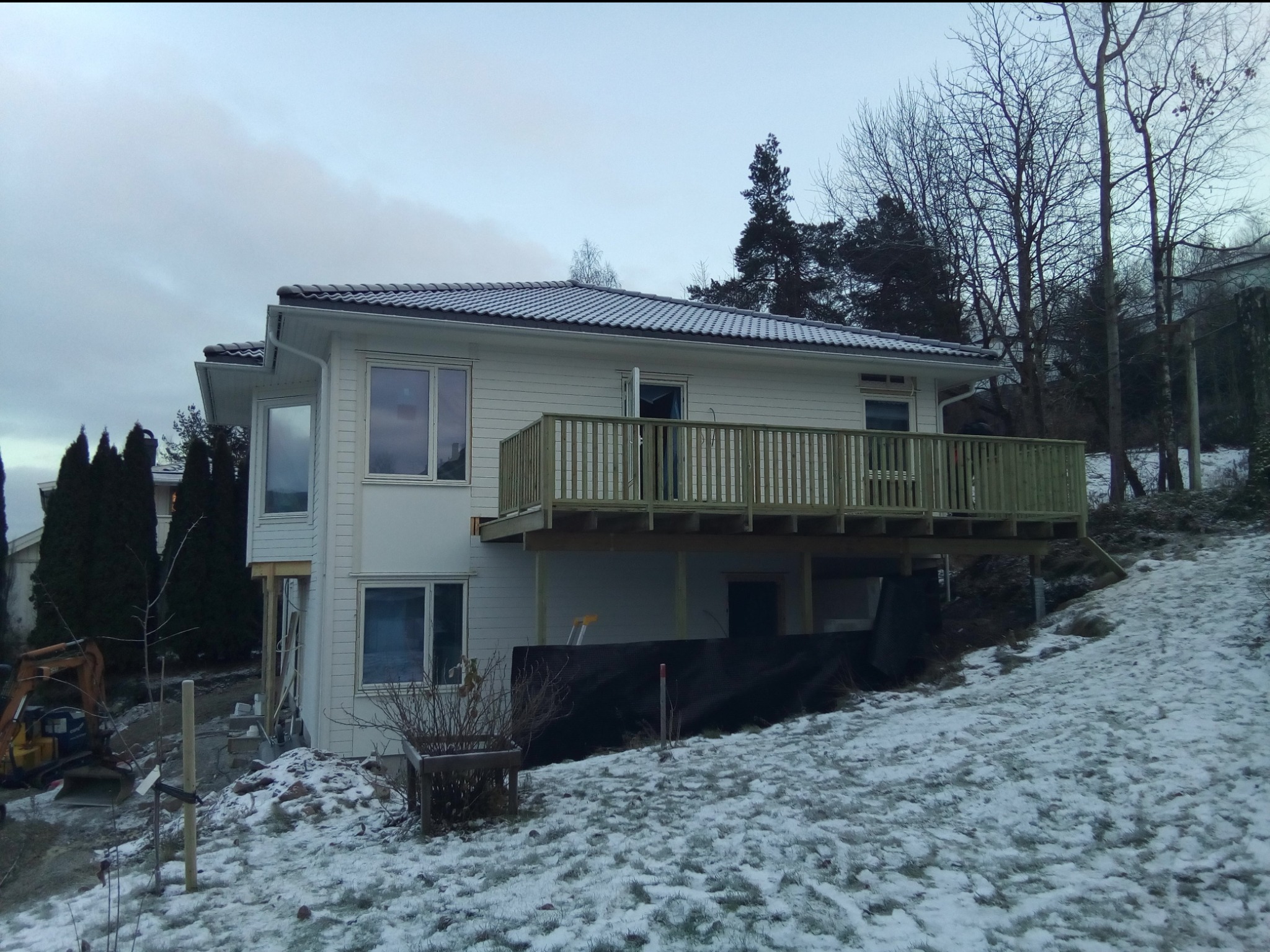Dwukondygnacyjny dom z jasną elewacją i drewnianym balkonem w trakcie budowy, otoczony śniegiem i drzewami, z widocznym mini-koparką i materiałami budowlanymi.
