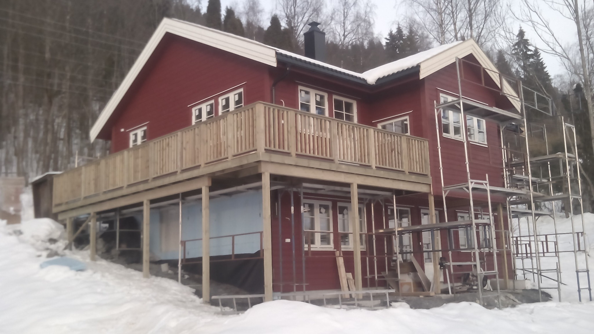 Czerwony dom z drewnianym balkonem na podwyższeniu, w trakcie budowy lub wykończenia, otoczony śniegiem, widoczne rusztowania i materiały budowlane.