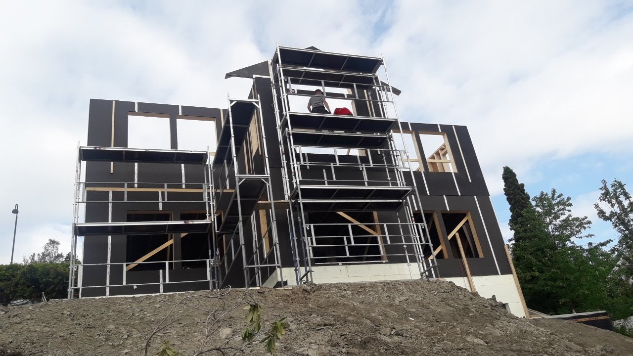Wznoszenie konstrukcji domu z czarną elewacją, widoczne rusztowania i pracownik na górnym poziomie, całość na tle pochmurnego nieba.