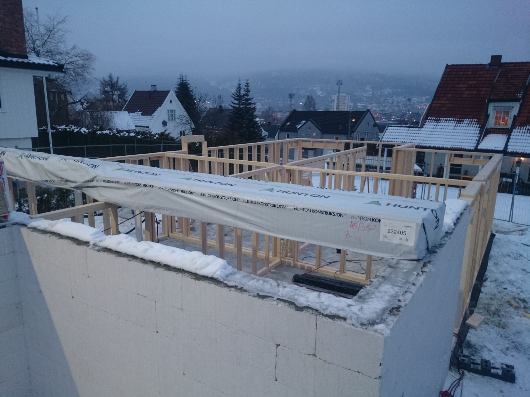 Wznoszenie konstrukcji domu szkieletowego na fundamencie z bloczków betonowych w zimowej scenerii, widoczne elementy drewniane i opakowane płyty Hunton Konstruksjon.